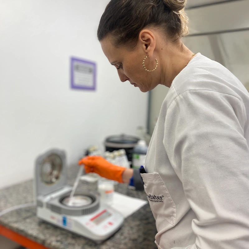 Mulher usando jaleco branco com o logo da Madater, manipulando uma amostra em um equipamento de laboratório.