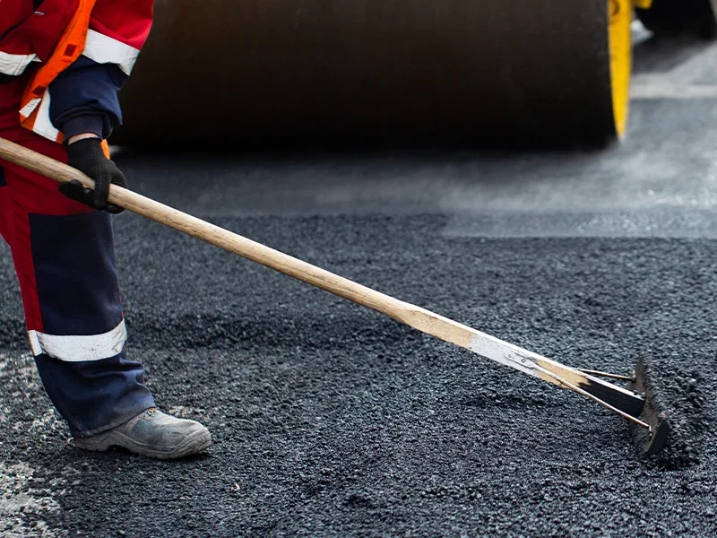 Trabalhador da construção civil ajustando o asfalto com uma ferramenta manual, enquanto um rolo compactador está visível ao fundo. O trabalhador usa uniforme de segurança, com colete laranja e calças com faixas refletivas. O foco está na preparação da superfície de asfalto, parte do processo de pavimentação de estradas ou ruas.