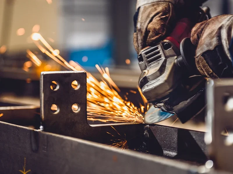 Trabalhador utilizando uma ferramenta de corte para metal, com a lâmina girando rapidamente enquanto gera faíscas. Ele usa luvas de proteção, e o foco é na precisão do trabalho de acabamento em uma peça de metal, com detalhes da superfície e os cortes sendo realizados.