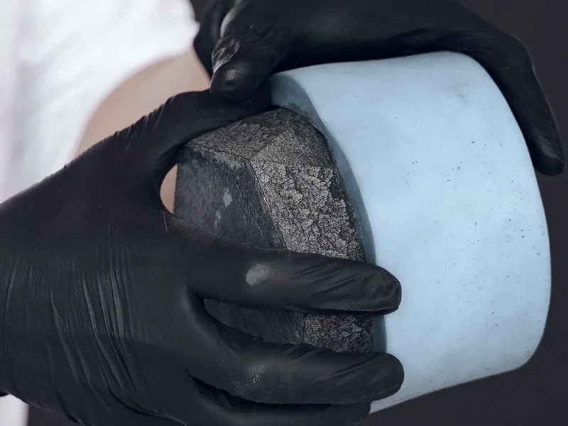 Uma pessoa usa luvas pretas enquanto segura dois materiais relacionados à fabricação de moldes: um objeto azul claro (silicone) utilizado para moldagem, e um objeto preto, que pode representar uma peça rígida ou já moldada.
