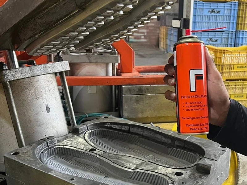 Mão segurando spray desmoldante ao lado de molde metálico com formato de solado, em ambiente industrial com máquinas de injeção e caixas plásticas ao fundo.
